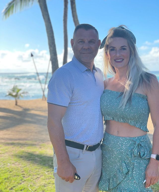 Mark Selman in a white t-shirt and pants posing with his wife in a green top and beautiful designer skirt.